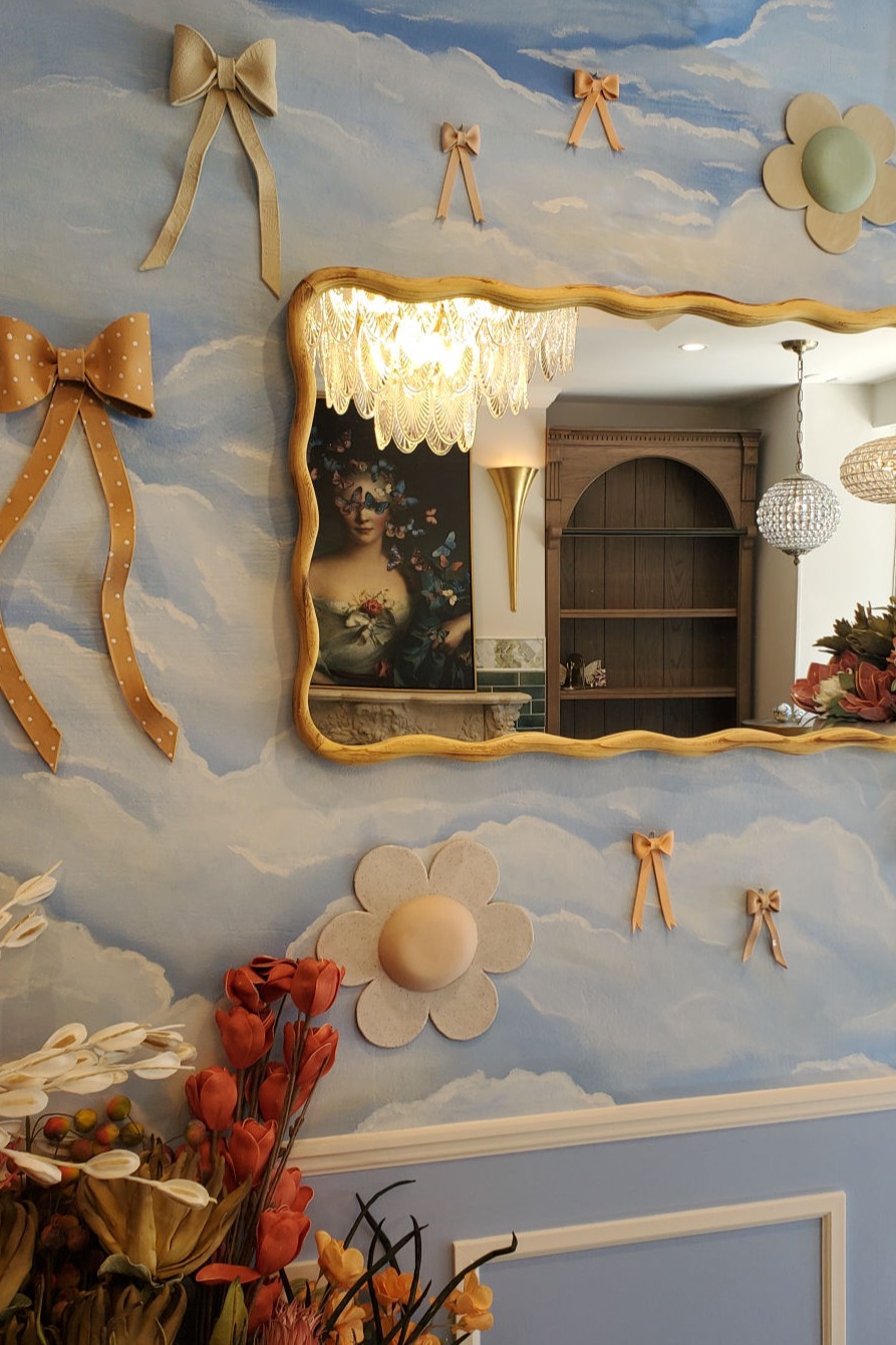 Cloud-painted blue wall decorated with beige bows, flowers, and a wavy gold mirror